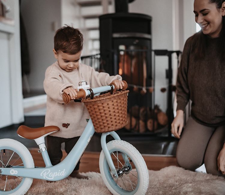 The 2-year-old boy is playing with a balance bike in the living room, accompanied by his mother who is nearby.