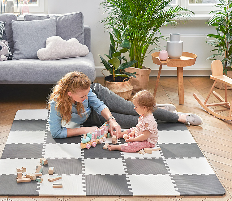 The one-year-old girl is playing with her mom on the Kinderkraft foam mat in the living room. They are building a train with wooden blocks.