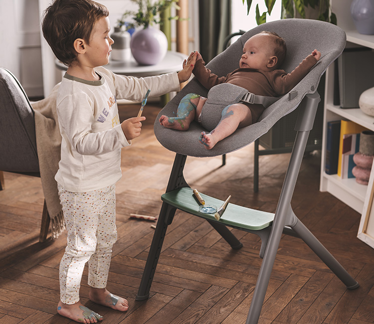 A two-year-old boy is enjoying some creative playtime with his younger brother, both happily engaging in finger painting with brushes. The younger sibling, a baby boy, is comfortably seated in the LIVY and CALMEE baby bouncer by Kinderkraft.