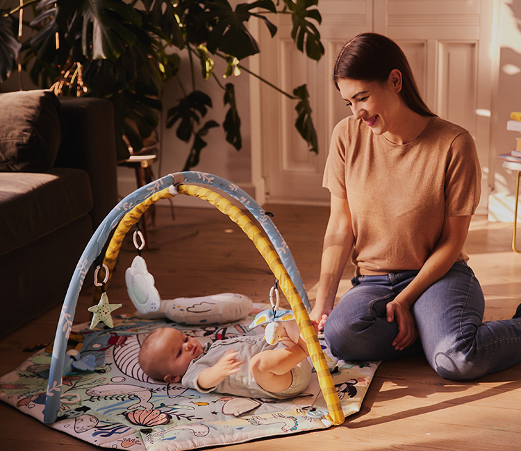 The baby is lying on their back on the play mat by Kinderkraft in the living room. Beside them, the mother is sitting, smiling at the baby.