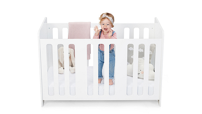 A Kinderkraft cot with rungs stands on a white background. There is a laughing little girl in the cot with mascots scattered all around her.