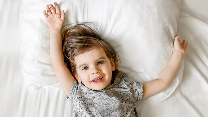 A few-year-old boy is lying on his back in bed on white sheets. He looks into the lens and spreads his hands to the sides, showing that he is comfortable.