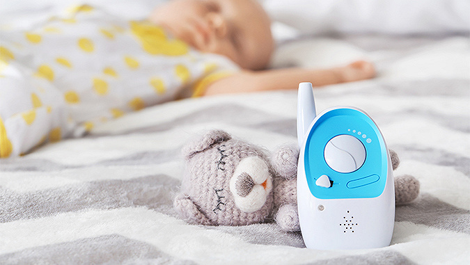 On the bed there is a white and blue Kinderkraft baby monitor leaning on a woollen mascot - a teddy bear. In the background, a child is sleeping on the same bed.