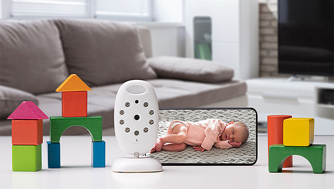 On the table there are houses arranged from blocks, and next to it, a Kinderkraft baby monitor and a smartphone displaying an image of a sleeping child.