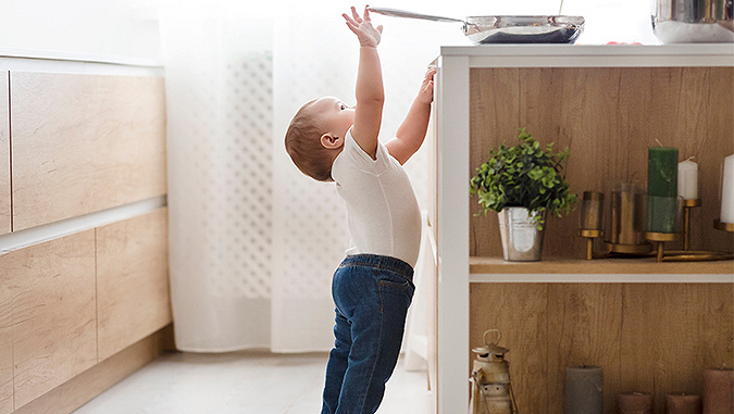 A little child wearing jeans and a white blouse is standing in the kitchen. The child is standing on its toes and reaches to the pan on the table.