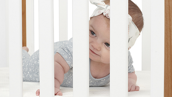 A few-month-old baby lying on a comfortable mattress looks through the white rungs of the Kinderkraft cot.
