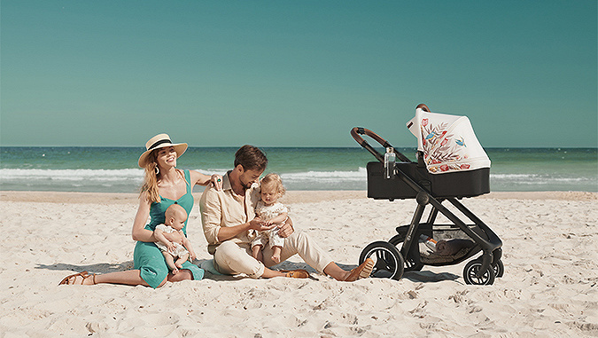 On a sunny day, parents sit on the seaside with their two young children. Next to them there is a Kinderkraft carrycot with a colourful hood.