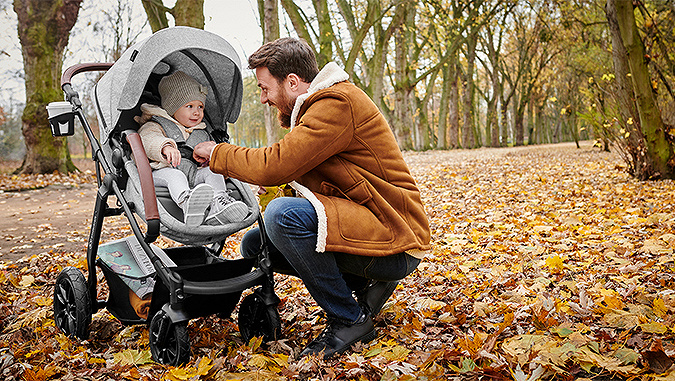 In a park full of autumn leaves, a laughing dad bends over a Kinderkraft stroller and adjusts the girl's safety harness.
