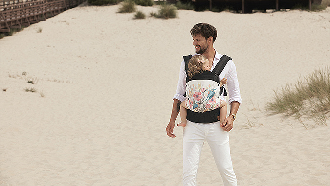 An elegantly dressed dad in a white shirt and trousers is standing on the beach. A little girl is sitting in a Kinderkraft carrier on his front.