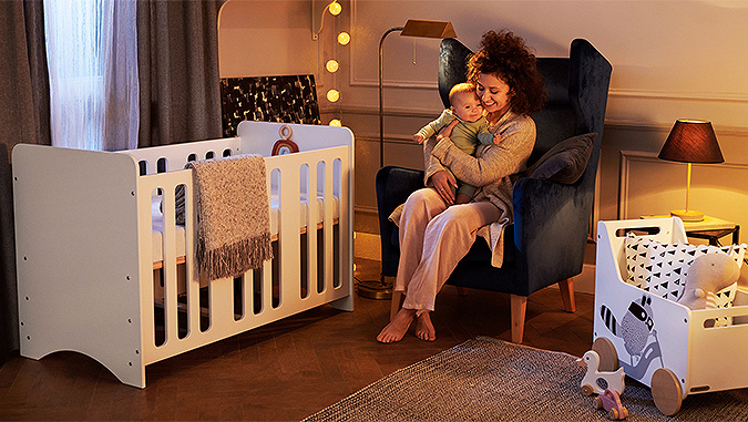 In the evening, in the children's room, mum holds a little child while sitting in an armchair. Next to them there is a playpen and a Kinderkraft Racoon toy chest.