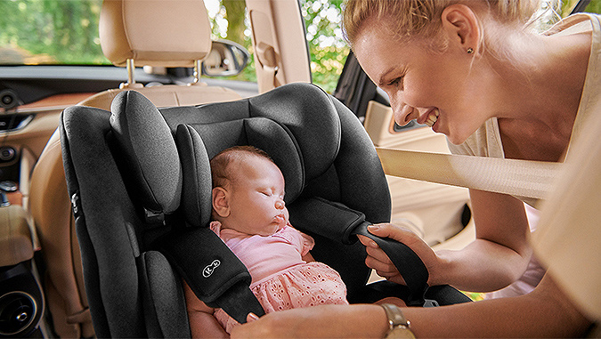 New-born baby sleeps in a grey Kinderkraft car seat placed forward facing in the car, and the smiling mother fastens the safety harness.