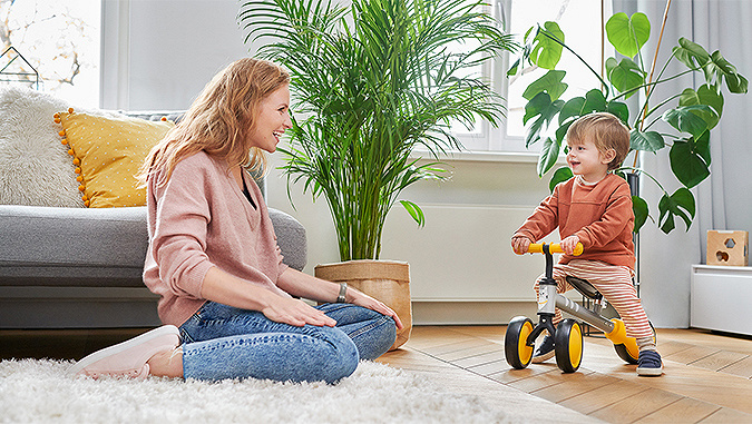 A little boy is riding a Kinderkraft tricycle around the room. His laughing mother, who is sitting on the floor next to him, is watching him.