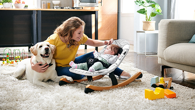 Mom sits at home on the carpet and caresses her little baby lying in a Kinderkraft car seat.  She hugs the dog with her other hand.