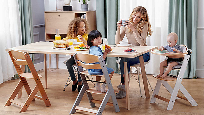 Mom and her three children eat breakfast sitting at the table. Two younger children sit on special Kinderkraft chairs.