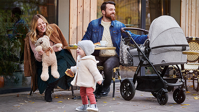 Laughing family at the café outdoors. Mom shows the girl the mascot, and the father looks at the other child lying in a Kinderkraft pushchair. 