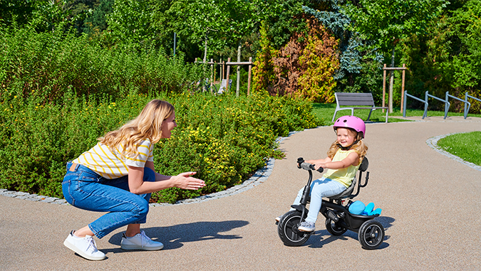 On a path in the park on a sunny day, a girl in a pink helmet rides a Kinderkraft tricycle towards a smiling mother.