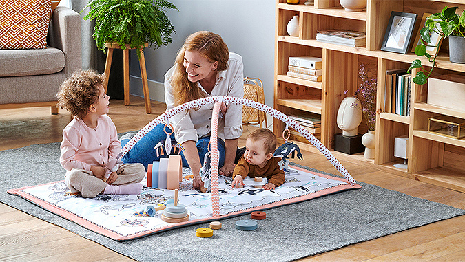 A Kinderkraft play mat is placed on the carpet in the apartment. Mother, a few-year-old child and a baby play on it.