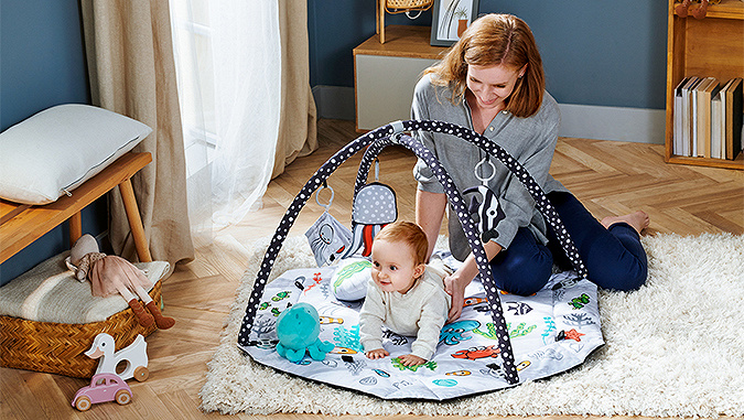 The Kinderkraft play mat is placed in the room on the carpet. Mother is sitting on the ground and is supporting a little baby lying on it.