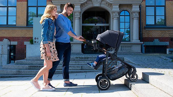 On a sunny day, smiling parents walk with a child sitting in a Kinderkraft stroller. The hood protects the baby from the sun.
