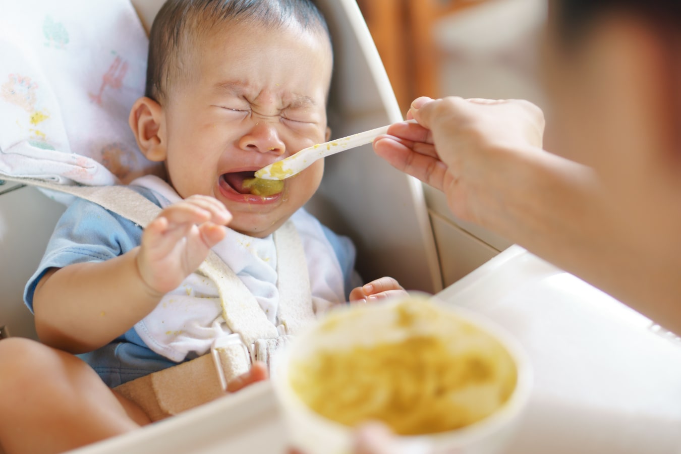The child sits fastened in a feeding chair, refuses to eat from the spoon, and cries.