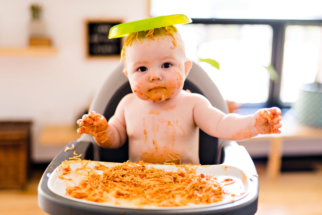 Dirty from spaghetti child, sits in a feeding chair, has a plate with food on the head.
