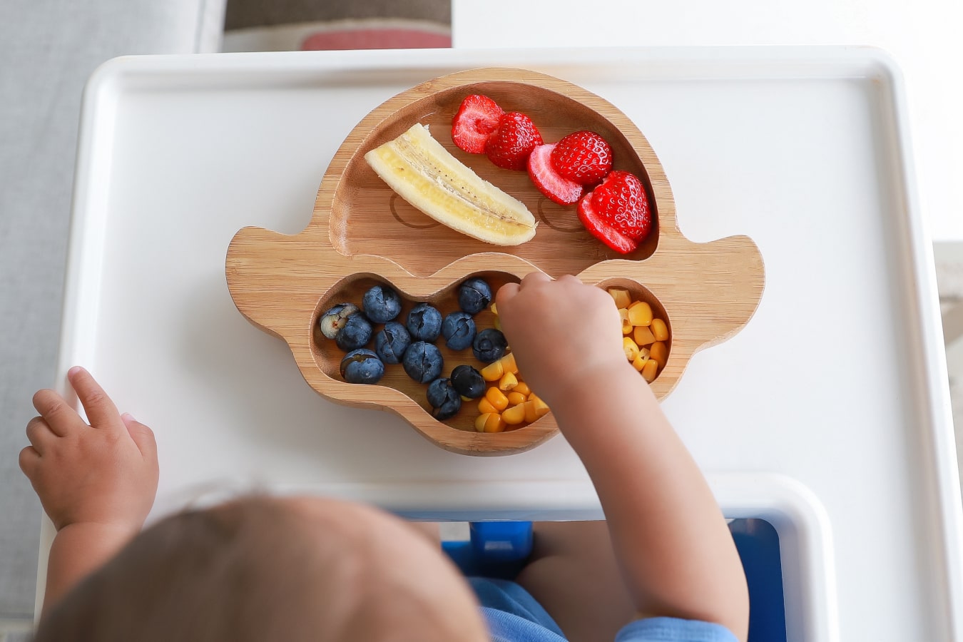 On a wooden plate with compartments colourful fruit and vegetables: banana, blueberries, corn, and strawberries can be seen. The child reaches for the food.