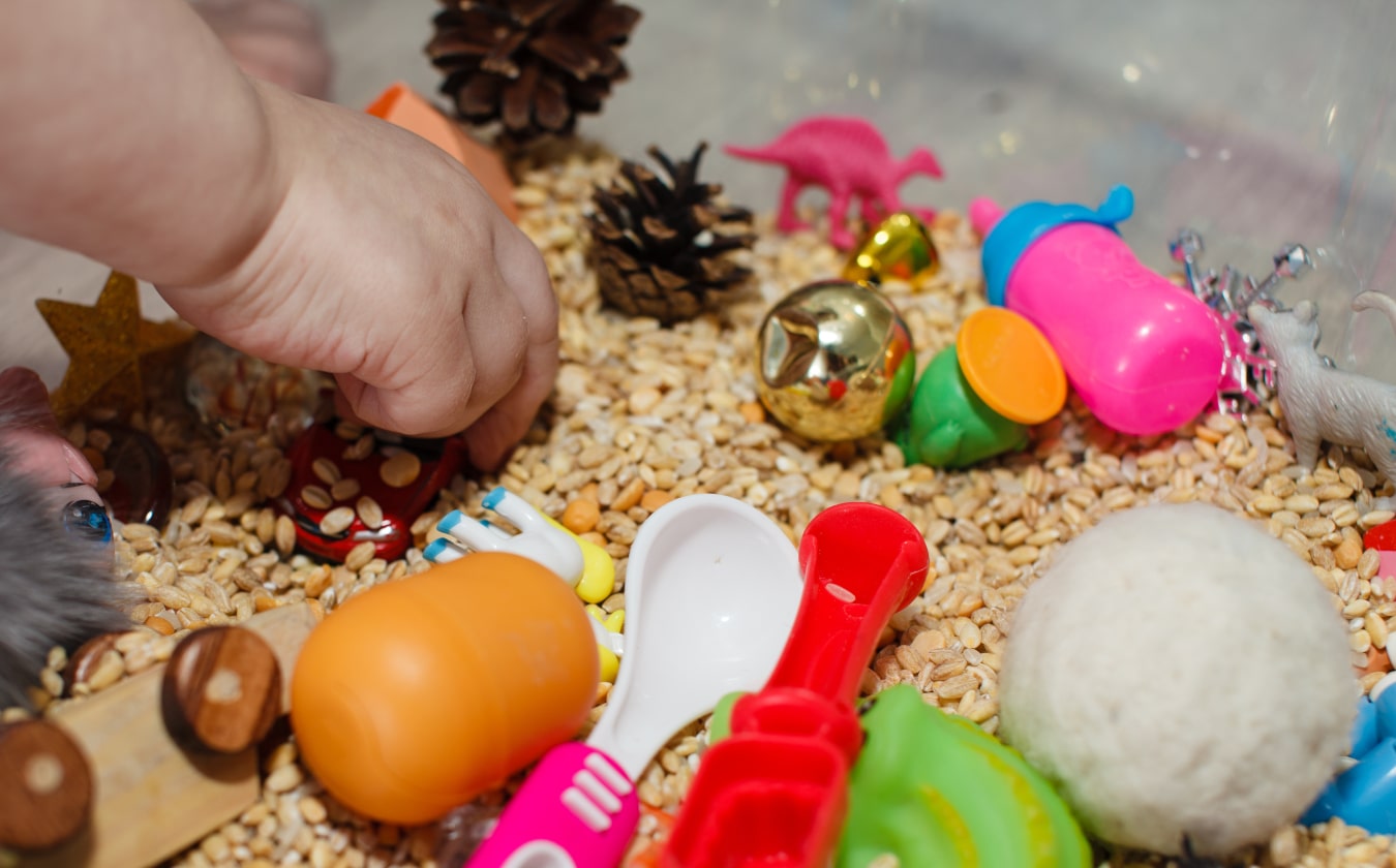 A child's hand touching grains, on which there are colourful children's toys. The child is learning new textures through touch