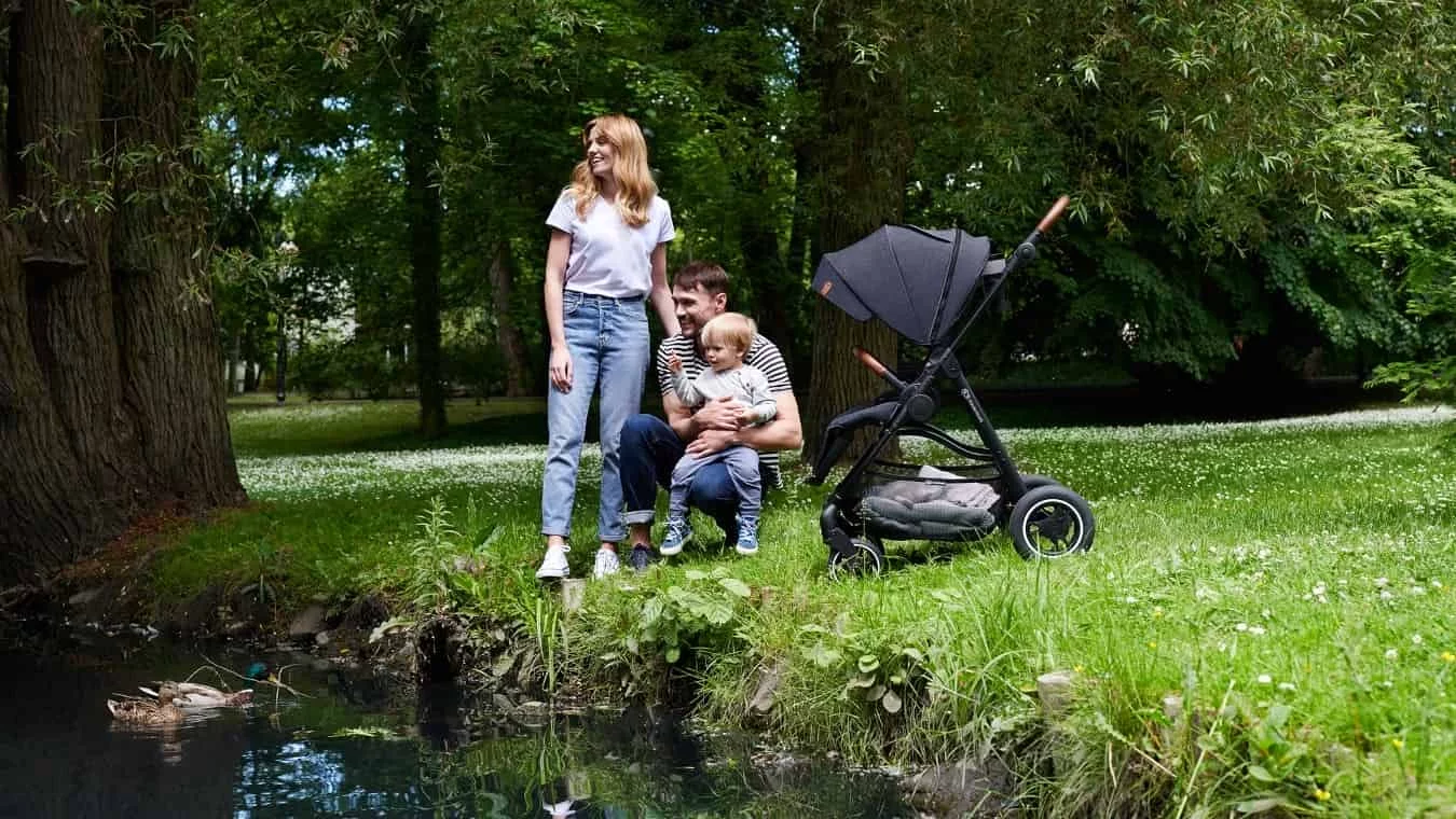 Mom and Dad are laughing. It's spring time, dad is holding his son in his lap, next to them there is the Kinderkraft ALLROAD stroller