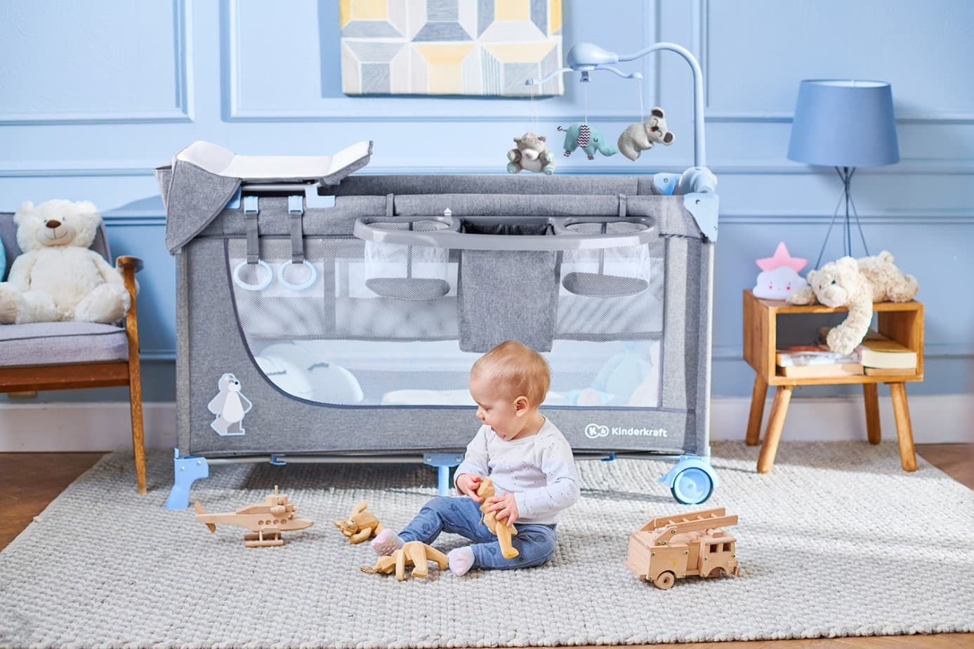 A small child playing on the carpet with wooden toys next to the JOY travel cot in the blue children's room