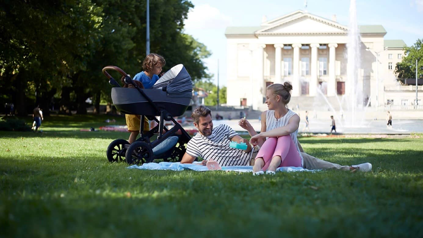 The family with mum and dad are sitting on a blanket in the grass in a city park, the son is standing next to a pram