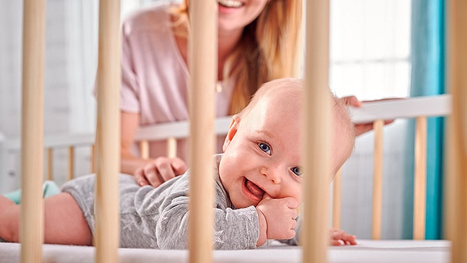 Smiling baby lying on his tummy in a wooden crib with rungs with his mother in the background behind him