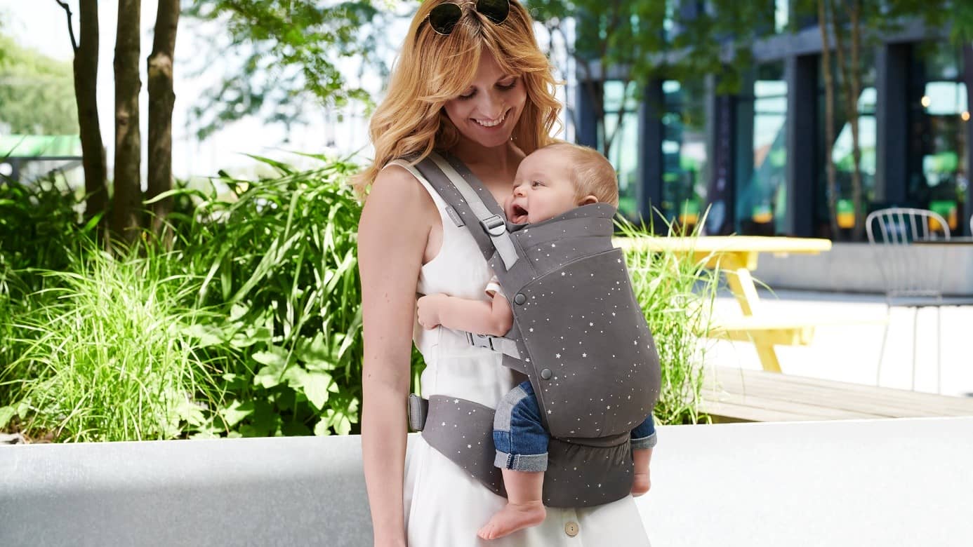 Mum carries the baby in the front in a HUGGY carrier outside while standing next to the plants
