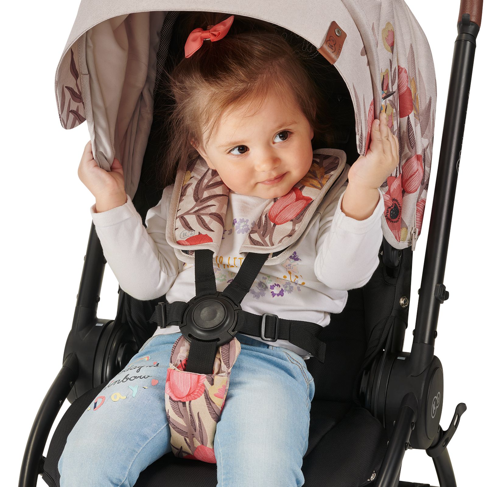 A little girl is sitting down in a stroller and is secured with a safety harness – she's grabbing the sun shade and smiling