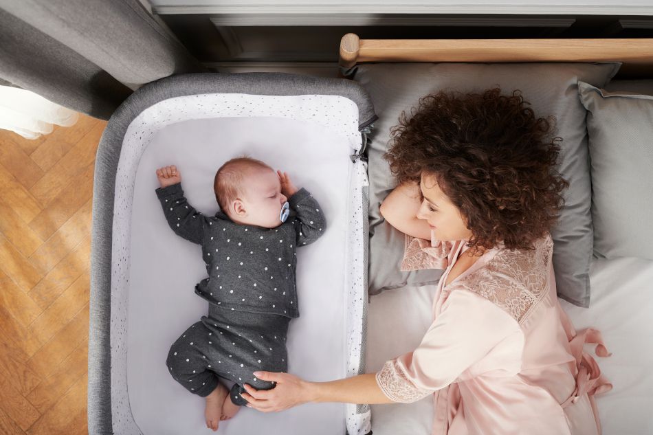A dark-haired woman is lying on a bed, with a child sleeping next to her in a co-sleeper cot. The woman is smiling, and the child is sleeping peacefully in a long-sleeved bodysuit.
