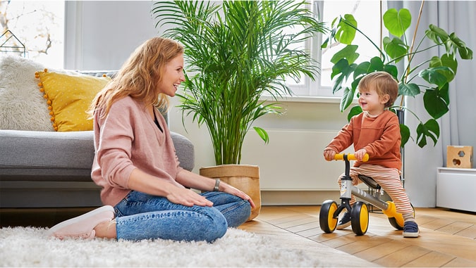 Mom laughs at a child riding a cutie kinderkraft tricycle. They are in a house, you can see green plants and a grey couch in the background. The child is smiling.